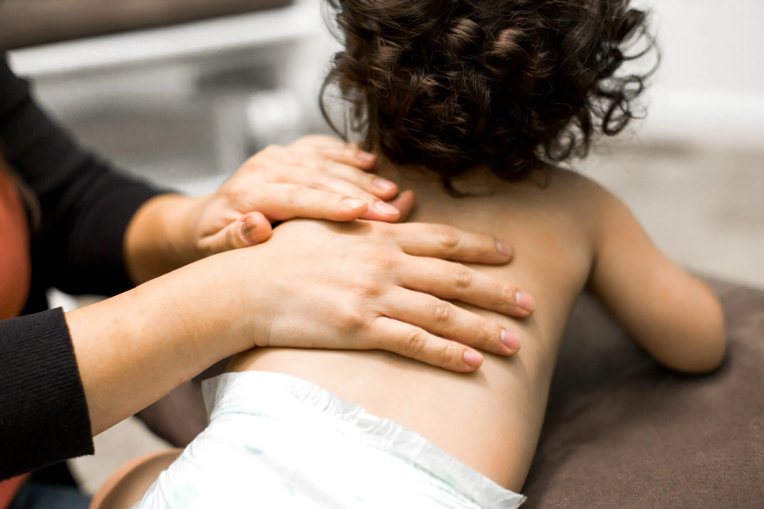 An adult is massaging the back of a toddler wearing a diaper on a cushioned surface in a softly lit indoor setting.