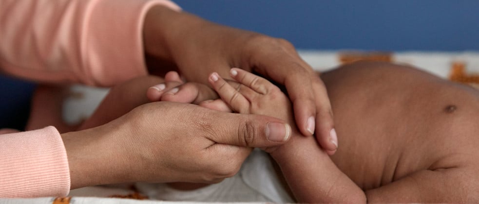 Hands gently holding a baby’s feet; the baby is lying on a patterned cloth with a blue background.