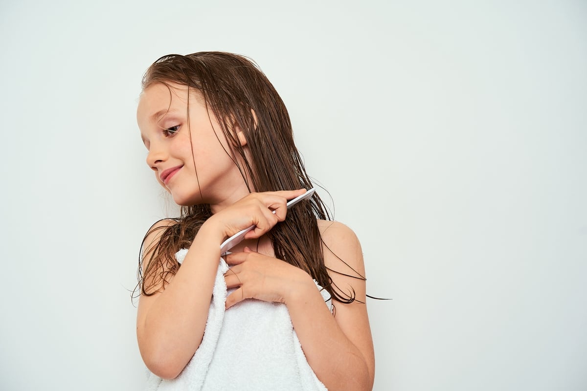 Little girl with wet hair wrapped in a towel, holding a comb and smiling slightly to the side.