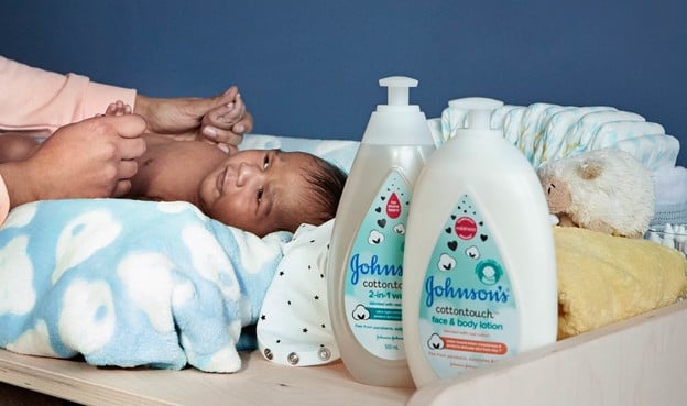 Baby lying on a changing table getting cared for by an adult with Johnson's baby products and diapers nearby. Text on bottles: "Johnson’s Cottontouch 2-in-1 Wash" and "Johnson’s Cottontouch face & body lotion."