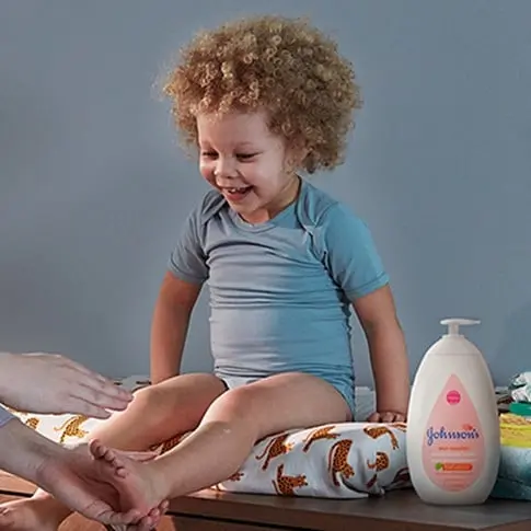 A happy baby with curly hair sitting on a changing table, wearing a blue onesie, with a parent’s hand reaching towards them and a bottle of Johnson's baby lotion nearby.