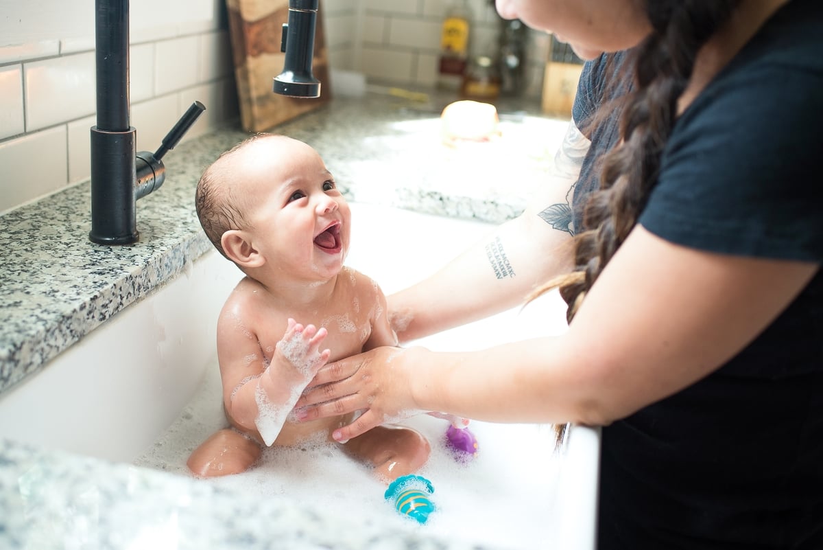 Madre bañando a un bebé sonriente en el fregadero de la cocina, rodeado de burbujas y juguetes.