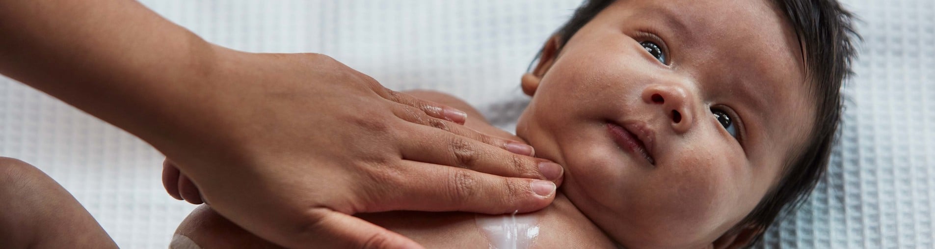 An infant is lying on a white, textured surface, while an adult hand gently massages lotion or oil onto the baby's chest.