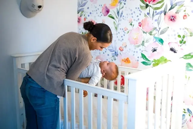 A woman leans over a white crib, smiling at a baby inside. The room features a colorful floral mural and a decorative animal head on the wall.
