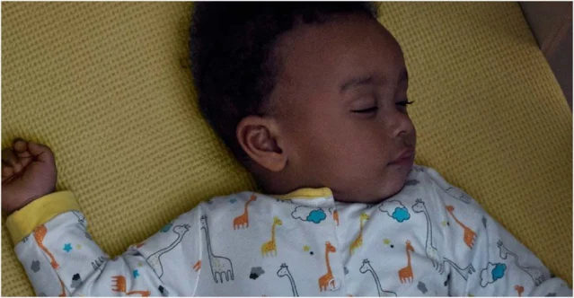 A sleeping baby lies on a yellow mattress, wearing white pajamas printed with colorful giraffes, clouds, trees, and stars.