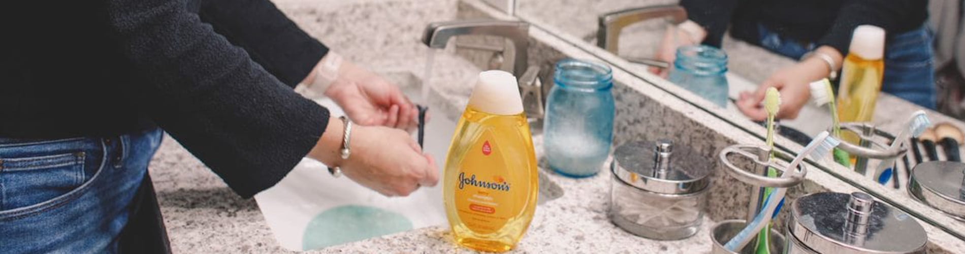 Woman washing her hands at a bathroom sink with various toiletries, including "Johnson's" baby shampoo, brushes, and containers, on a granite countertop.