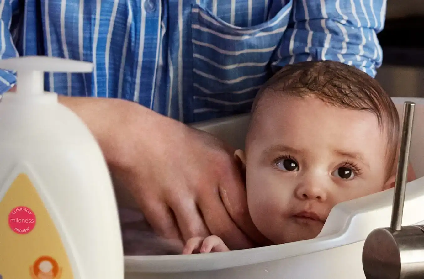 A baby is being bathed in a white tub by an adult wearing a blue striped shirt. A bottle with a label "Clinically mildness proven" is in the foreground.