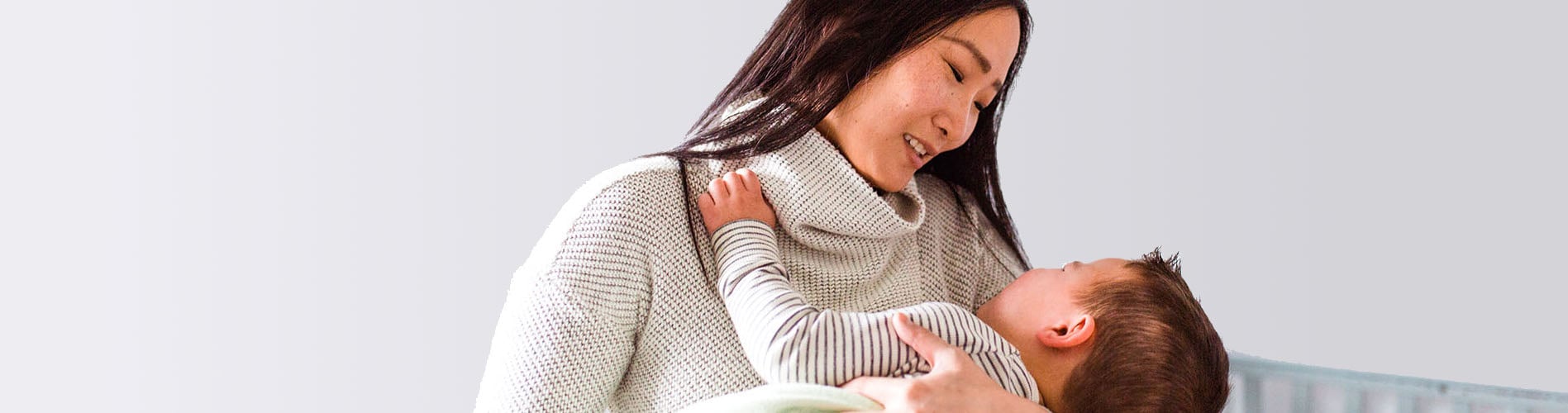 A woman holding and looking lovingly at a baby, dressed in a striped shirt, in a cozy environment with a light gray background.