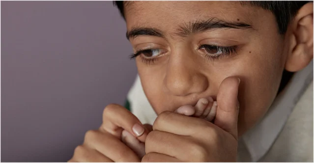 A child is thoughtfully biting their fingernail in a neutral-colored room, their hands close to their face, staring intently at something out of the frame.