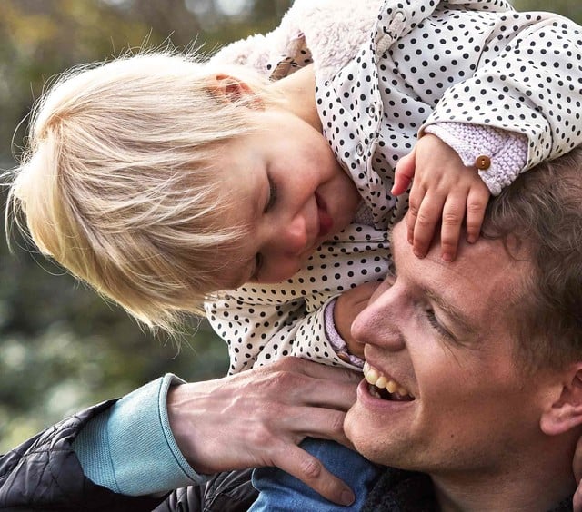 Un hombre lleva sobre sus hombros a un niño sonriente que viste una chaqueta a lunares, con vegetación exterior de fondo.