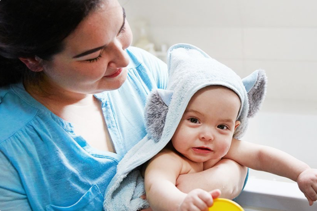 Smiling baby in a blue towel with bear ears being held by a woman in a blue shirt, in a bathroom setting.