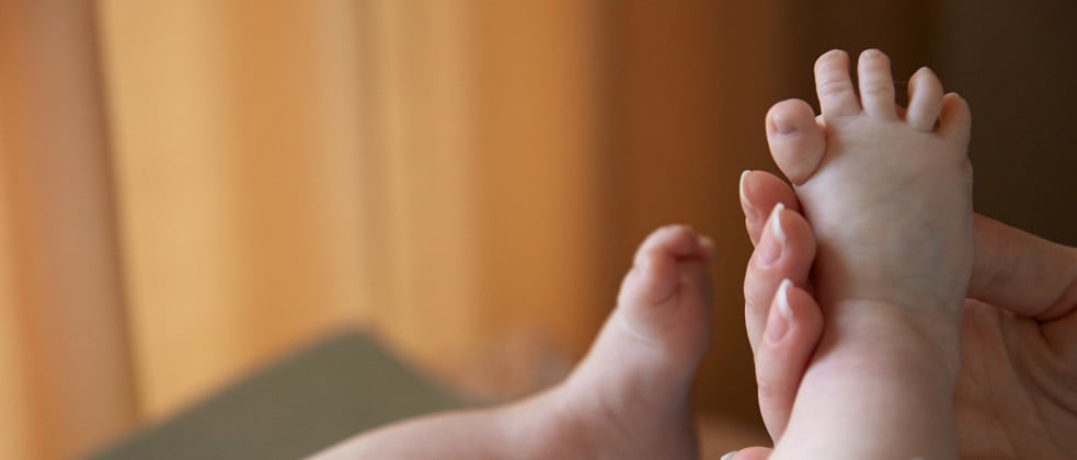 Baby’s feet are gently held by an adult hand, with a blurred, softly lit, warm-toned background.