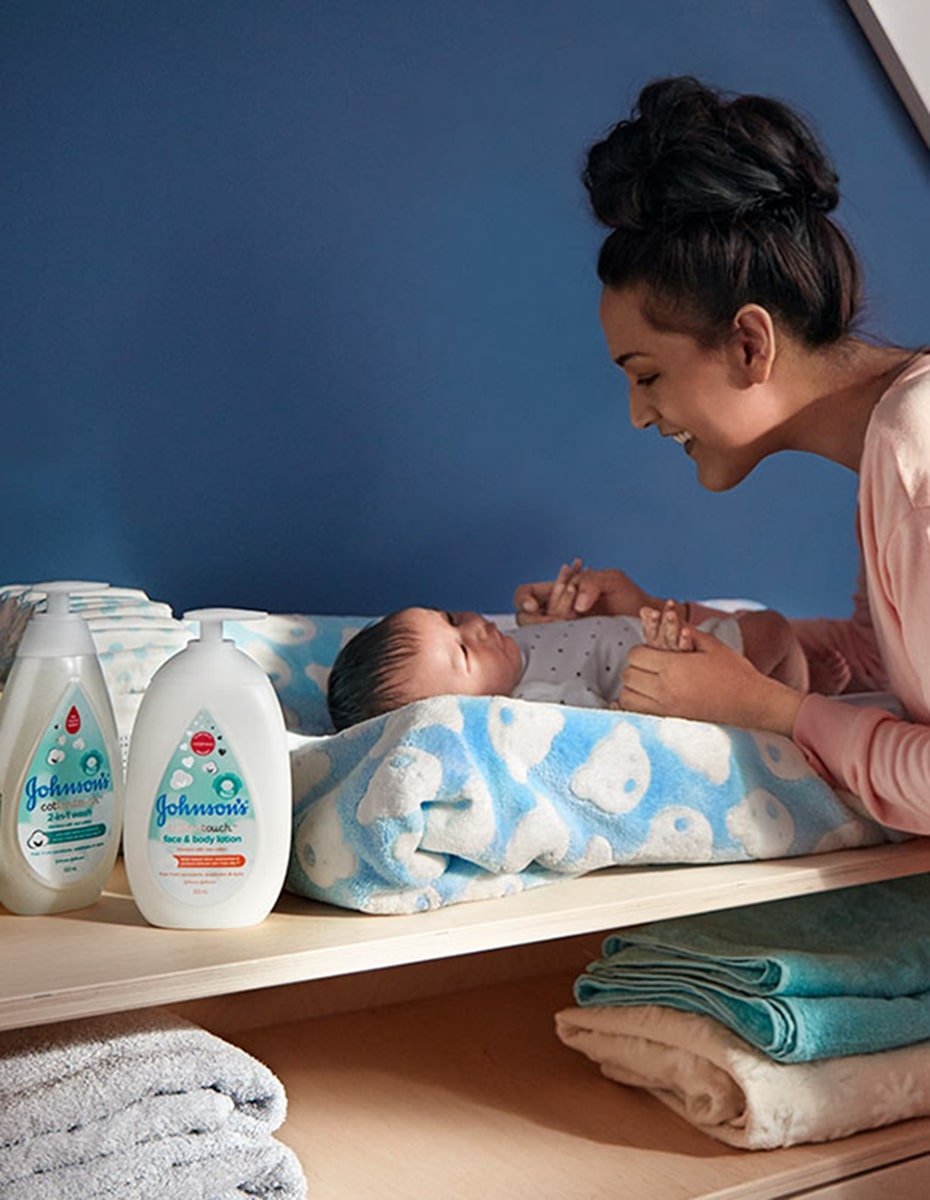 Mother smiling at her baby lying on a changing table with blue patterned towel, next to Johnsons baby bath products.