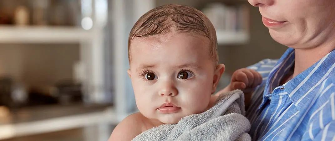 A baby wrapped in a gray towel is held by a person in blue-striped pajamas, in a room with blurred shelves in the background.