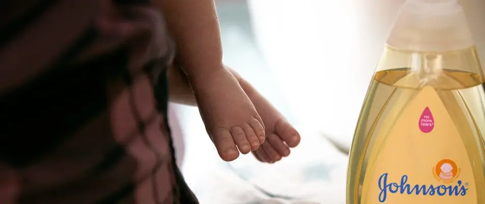 A close-up shows a baby's feet while being held, with a bottle of Johnson's baby shampoo featuring the text "no more tears" next to it, set in a softly lit room.
