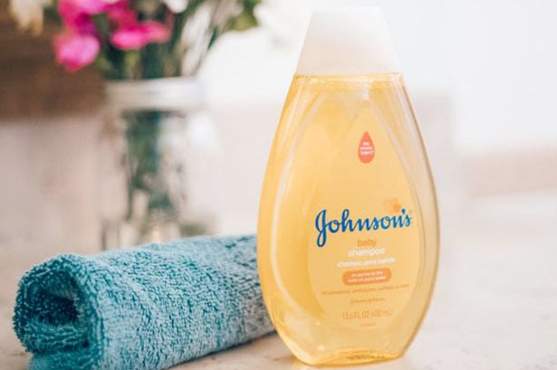 Johnsons Baby shampoo, 13.6 fl oz (400 ml) yellow bottle with a white cap, placed on a countertop next to a blue towel, with flowers in the background.