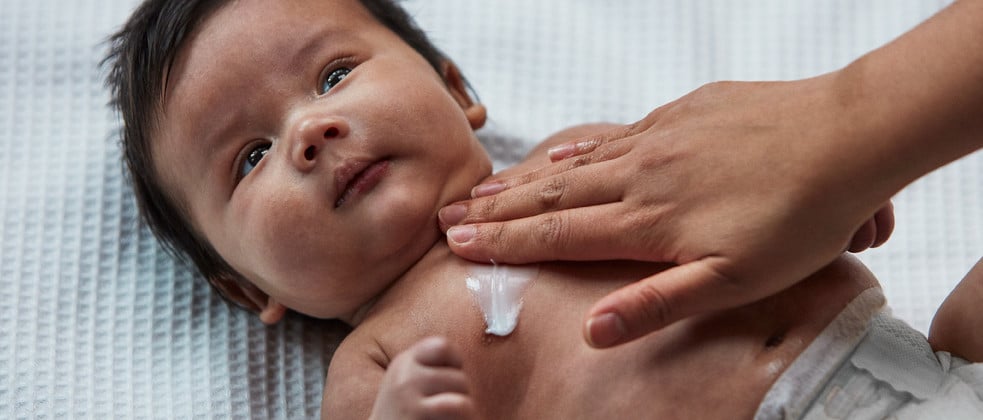 A baby with dark hair lies on a white, textured surface while an adult hand applies lotion to the baby's chest.