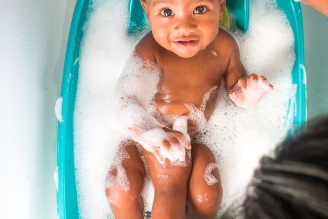 A baby smiles while sitting in a teal bathtub filled with bubbles, with a parent partially visible at the bottom of the image.