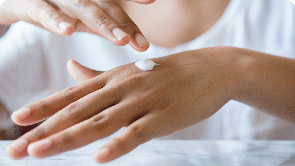 A hand applying a small amount of white lotion to the back of the other hand, with a marbled surface and a blurred white shirt in the background.