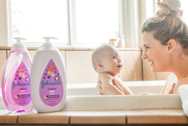 Una madre baña a su bebé sonriente en un baño iluminado por el sol con los envases de loción y jabón líquido de Johnson's Baby Bedtime en primer plano.