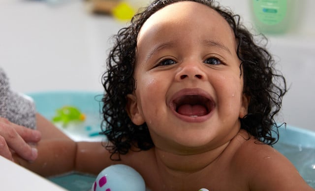 A baby with curly hair is smiling while playing in a bathtub, surrounded by bath toys and a person's arm, with bathroom items visible in the background.