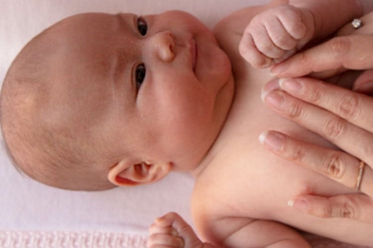 A baby lies on a pink blanket while an adult’s hands gently touch the baby's hands and torso, creating a tender moment.