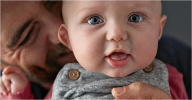 Baby with wide eyes and open mouth being held by an adult (partially visible) with a beard, in an indoor setting. The adult appears to be smiling warmly.