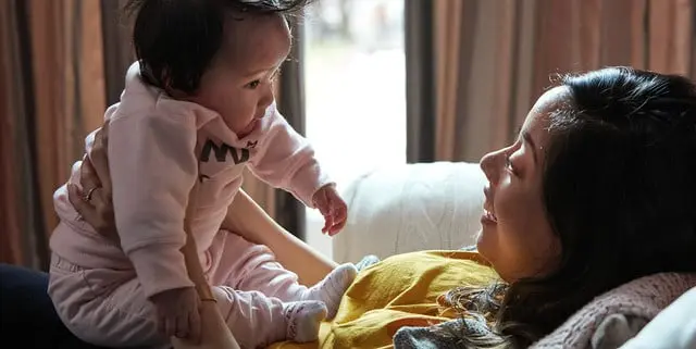 A woman lies on a couch holding a baby who is dressed in pink. They share a moment in a warmly lit room with curtains in the background.