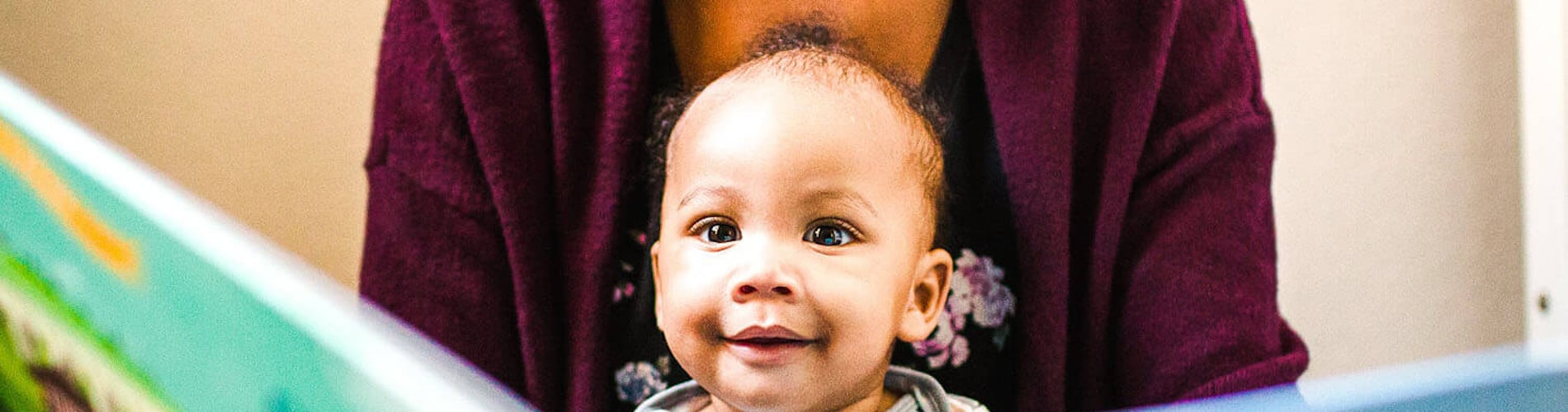 A smiling baby is seated on a person's lap while they read a colorful book together in a cozy, indoor setting.