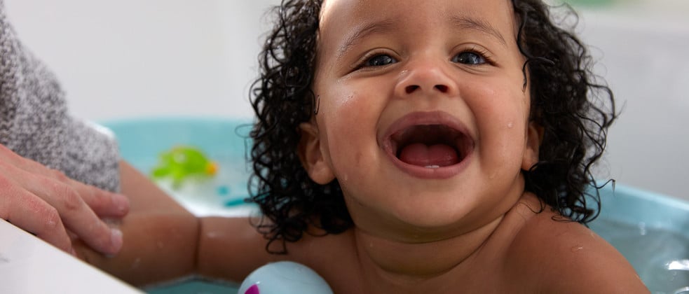 A baby with curly hair laughs while sitting in a bathtub filled with water, accompanied by a supportive adult hand. A small green toy floats in the background.