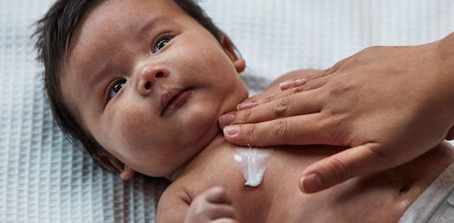 Baby lying down with lotion applied on chest, while a hand gently rubs the lotion in.