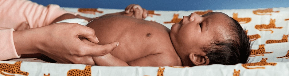 A parent holds a baby's hand while the baby lies on a cheetah-print changing pad, wearing a diaper and looking up.