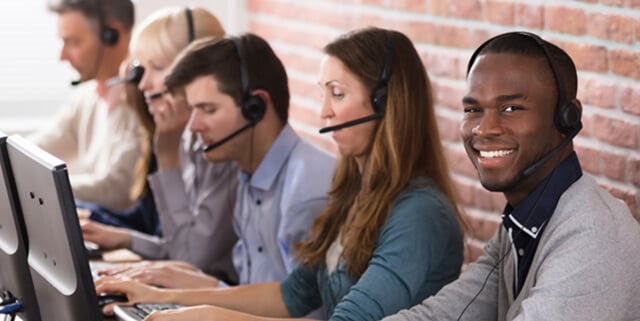 Customer service representatives at desks, wearing headsets and assisting callers, with one representative smiling at the camera.