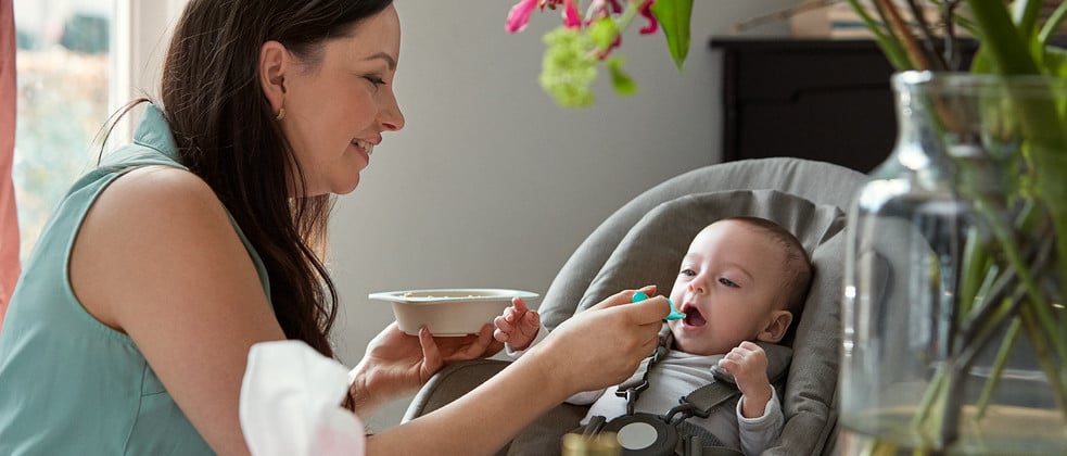 A woman feeds a baby with a spoon, holding a bowl in a modern, well-lit room with plants and furniture in the background.