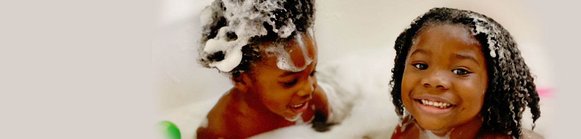 Two young children with curly hair having fun in a bubble bath, smiling and playing.