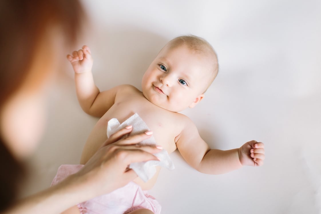 Bebé sonriente con pañal acostado boca arriba mientras su madre limpia con una toallita el pecho del bebé.