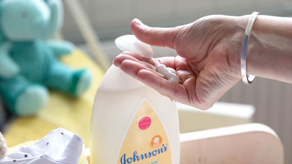 A hand dispenses baby lotion from a Johnson's bottle in a nursery, with a soft toy and baby clothing in the background. The bottle reads "Johnsons" and "Clinically Proven Mildness."