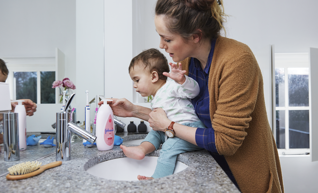 A parent helps a baby sit on a bathroom counter while holding a bottle of Johnson's baby lotion, with a sink and various items in the background.