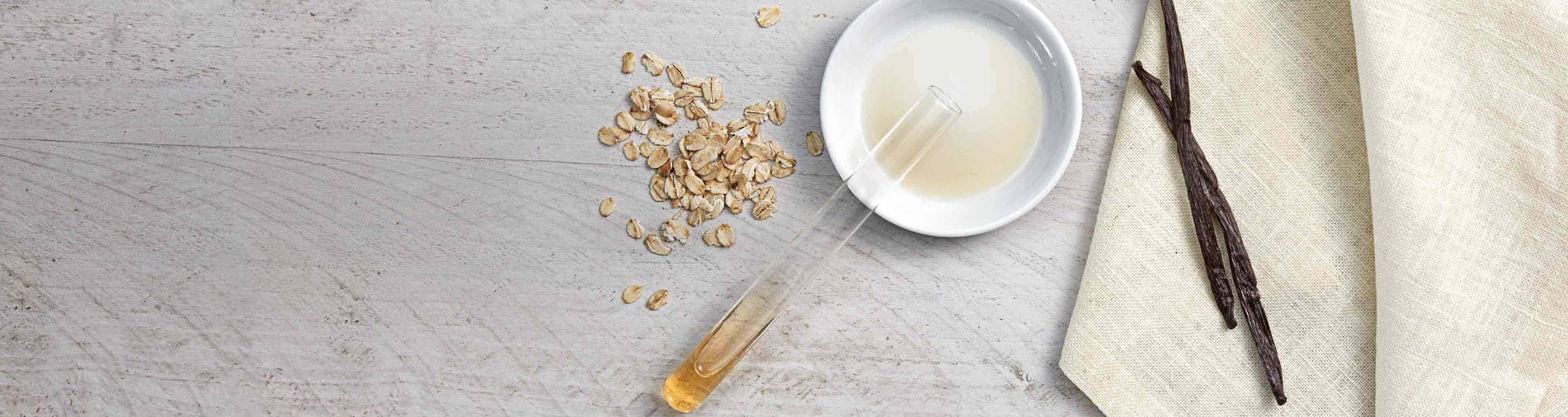 Scattered oat flakes and a test tube filled with liquid rest on a wooden surface, next to a white bowl containing liquid, vanilla beans, and a beige cloth.