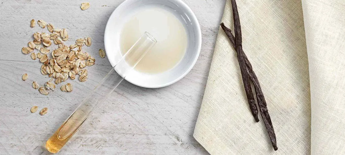 Scattered oat flakes and a test tube filled with liquid rest on a wooden surface, next to a white bowl containing liquid, vanilla beans, and a beige cloth.