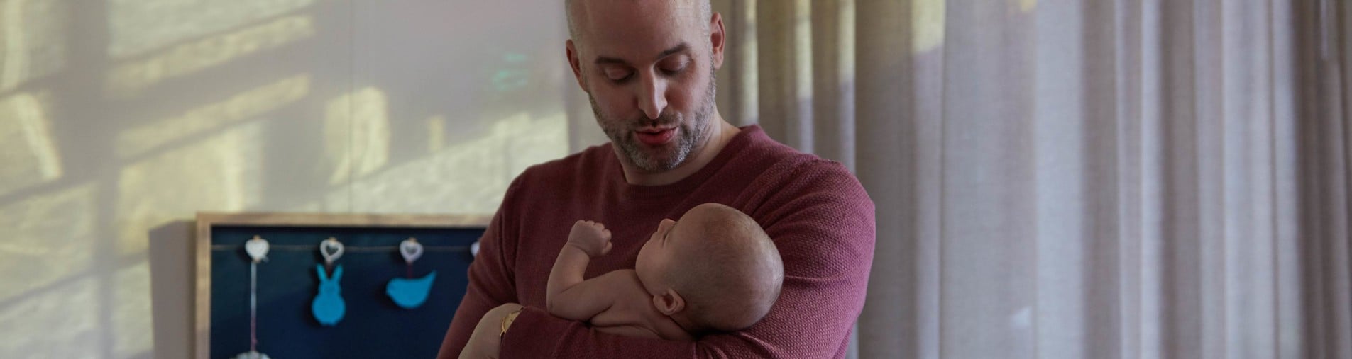 A person with a beard, in a red sweater, cradles an infant lovingly in a softly lit room with sheer curtains and a wall-mounted board with blue shapes hanging.