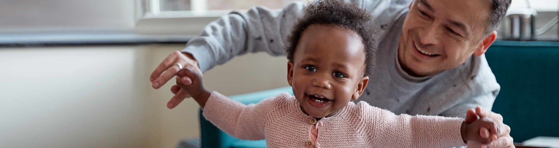 A joyful toddler, held up by an adult, smiles widely in a cozy, brightly lit living room near a window.