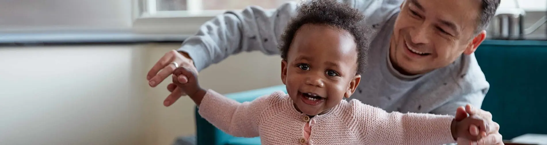 A joyful toddler, held up by an adult, smiles widely in a cozy, brightly lit living room near a window.