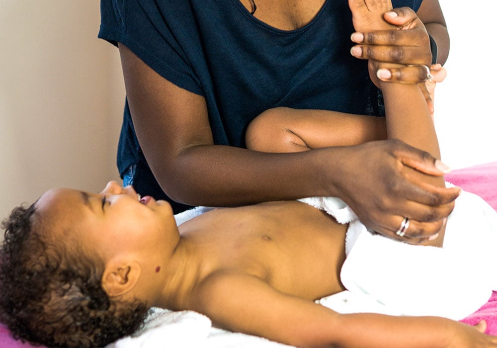 A child lies on a bed, partially wrapped in a white towel, while an adult gently stretches the child's arm; the background is a neutral-colored room.