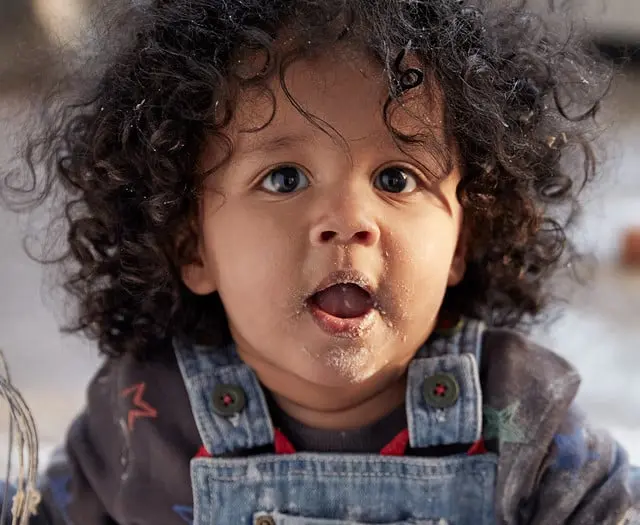 A young child with curly hair and food on their face looks directly at the camera, wearing a denim overall and a dark shirt with colorful patterns, indoors.