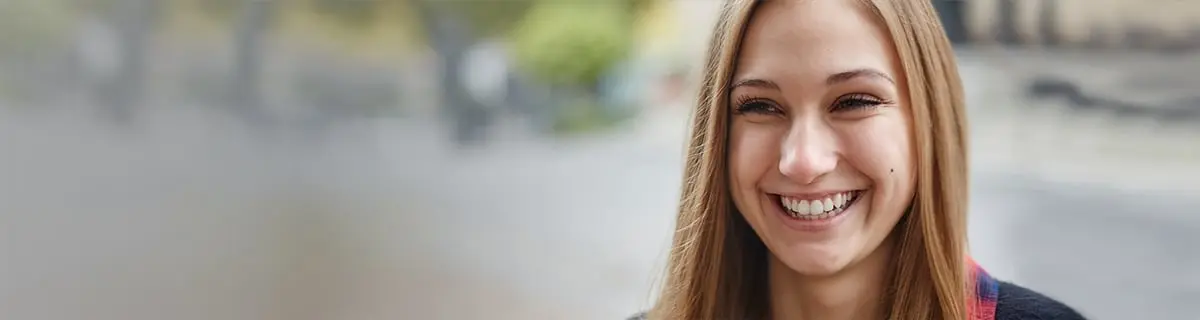 A woman smiles widely, her long hair falling around her face, in an outdoor setting with trees and blurred background, giving a cheerful and vibrant atmosphere.