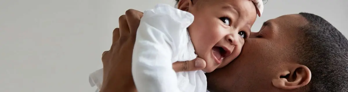 An adult holds a laughing baby close to their face, appearing to kiss the baby's cheek. The background is plain and light-colored.