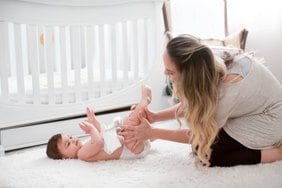 A mother playing with her baby on a white rug next to a crib in a bright nursery.