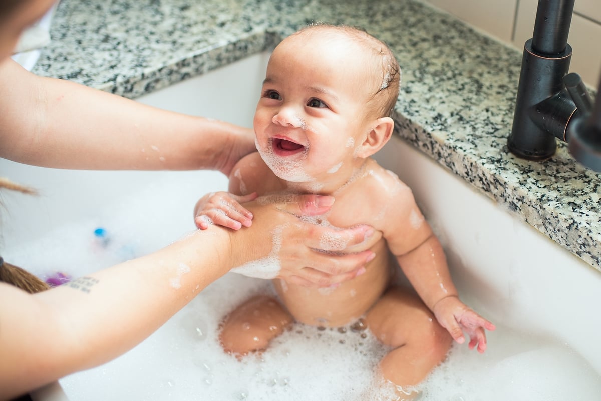 Smiling baby being bathed by their parent in a sink with foam.