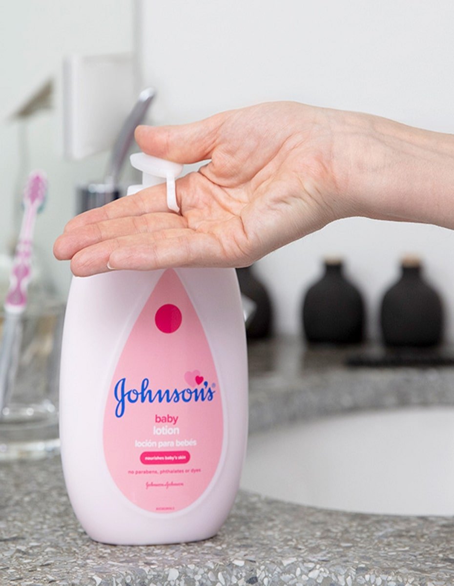 Hand pressing the pump of Johnsons Baby lotion, in a pink 13.6 fl oz (400 ml) bottle with text indicating clinically proven mildness and no parabens, phthalates, or dyes, on a bathroom countertop with toothbrush and bottles in the background.